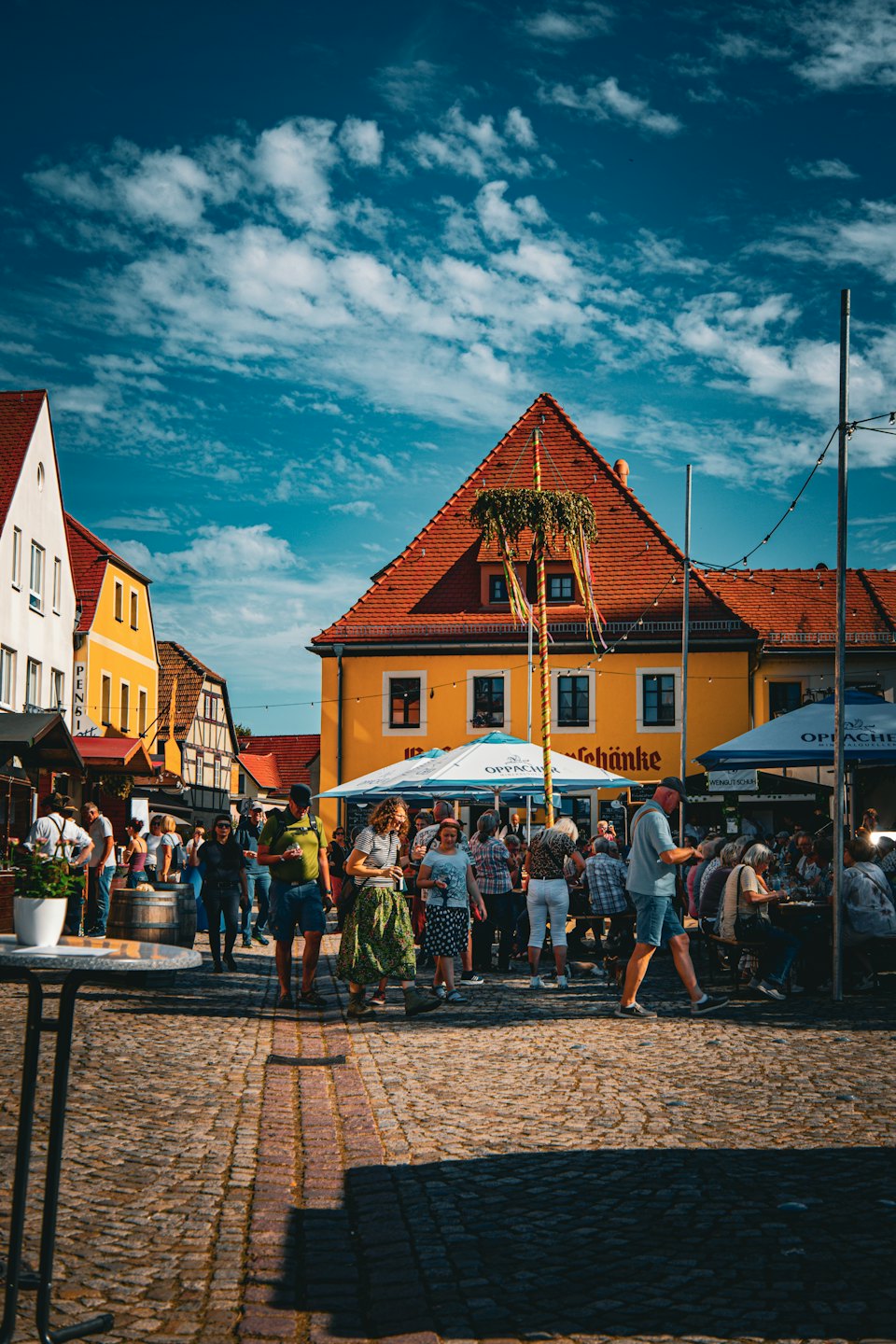 People socialize around a decorated Maypole in a town square.